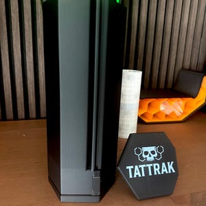 May include: A black, rectangular device with a vertical design, featuring a small, hexagonal emblem with the text "TATTRAK" and a skull graphic. The device is next to a green and black structure with colorful balls.