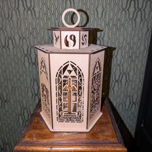 Könnte beinhalten: Eine lasergeschnittene Holzlaterne mit aufwendigen geometrischen und religiösen Designs. Die Laterne hat einen Griff, ein Kreuz und bogenförmige Ausschnitte. Sie steht auf einem Holztisch und zeigt ihre detaillierte Handwerkskunst und dekorative Ausstrahlung.