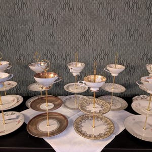 Könnte beinhalten: Mehrere mehrstöckige Servierstände mit Teetassen und Untertassen. Die Ständer haben goldfarbene Metallrahmen und halten Teller mit floralen und gemusterten Designs. Die Teetassen sind weiß mit goldenen Rändern. Der Hintergrund ist eine gemusterte Wand.