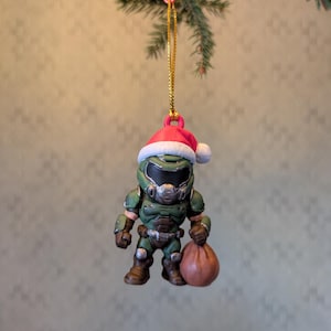 Könnte beinhalten: Ein Christbaumschmuck mit einer grünen und braunen Actionfigur, die einen Weihnachtsmannhut trägt und einen braunen Sack hält. Der Schmuck hängt an einer goldenen Schnur und einem rosa Stern.