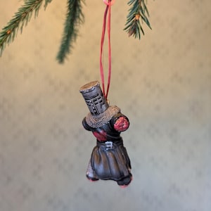 May include: A Christmas tree ornament shaped like a medieval knight. The ornament is primarily black with silver chainmail and red accents. It hangs from a red string and is suspended from a green pine branch.
