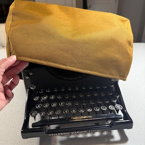 May include: A black Underwood Standard typewriter with a mustard-colored cover. The typewriter has a full keyboard with silver keys and a metal frame. The cover is rectangular and appears to be made of a canvas-like material.