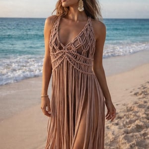 May include: A light brown macrame halter dress with fringe detailing. The dress has a plunging neckline and high side slits. The model is standing on a sandy beach with the ocean in the background. The dress is a bohemian style.
