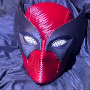 May include: A red and black mask with a textured surface. The mask features a red face and head covering with black accents around the eyes and ears. The eye openings have a white mesh material.