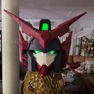 May include: A detailed, close-up view of a Gundam head sculpture. The head is primarily dark gray, red, and white, with glowing green eyes and a green light on top. The sculpture is mounted on a golden, petal-like base.