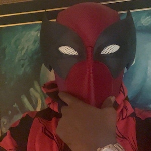 May include: A red and black full-head mask featuring large, white, perforated eye lenses and a textured fabric surface. The mask is paired with a red and black patterned hooded sweatshirt and a gold-toned wristwatch with a white face.