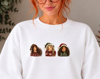 Può includere: Una felpa bianca a girocollo con una stampa grafica di tre figure illustrate che indossano cappelli e maglioni natalizi. Le figure hanno acconciature distinte e indossano abiti invernali con motivi rossi e verdi.