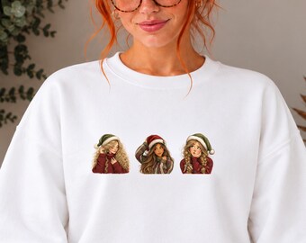 Può includere: Una felpa bianca a girocollo con una stampa grafica di tre figure illustrate che indossano cappelli da Babbo Natale rossi e verdi. Ogni figura ha un'acconciatura distinta e indossa un maglione rosso sul petto.