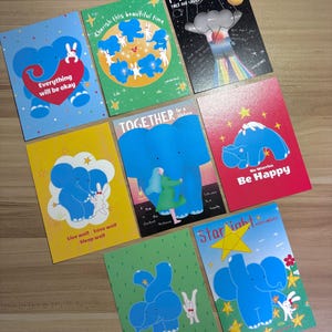 May include: A collection of colorful greeting cards featuring whimsical illustrations of blue elephants and white rabbits. Each card has a unique design with uplifting messages like "Everything will be okay" and "Be Happy."