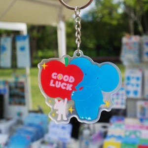 May include: A blue elephant keychain holding a red apple with the words "GOOD LUCK" printed on it. The keychain has a silver chain and ring. A small white figure is below the apple.