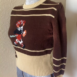 May include: Brown and beige striped sweater with a knitted design of a person wearing a kilt and a hat.