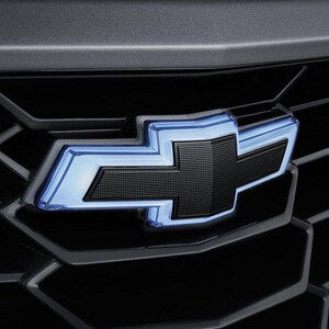 May include: Close-up of a black Chevrolet emblem with a blue illuminated outline, set against a dark gray background. The emblem is a stylized cross, with a textured surface.