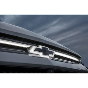May include: Close-up of a silver Chevrolet emblem on a dark gray vehicle. The emblem is a stylized cross shape with a black center. A chrome strip runs along the top of the vehicle, reflecting the cloudy sky.