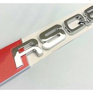 May include: A chrome-finished "RS Q8" emblem with a red and silver accent. The emblem is on a white backing, ready for application. The letters are bold and three-dimensional, reflecting light. The red accent is a rectangular shape.