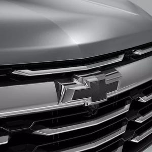 May include: Close-up of a silver car grille with a black Chevrolet emblem. The grille has horizontal chrome bars and a sleek, modern design. The hood of the car is visible above the grille.