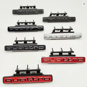 May include: A selection of Quattro car badges in black, silver, and red. Each badge displays the word "Quattro" in a bold font, mounted on a black plastic base. The badges are designed for automotive use.