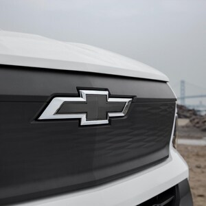 May include: Close-up of a white and black vehicle front end. The black grille features a silver Chevrolet emblem. The background shows a bridge and a pile of rocks under an overcast sky.