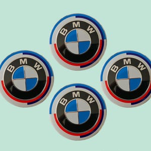 May include: Four circular BMW emblems with the BMW logo. Each emblem has a blue and white checkered design, a black ring, a red and white ring, and a blue outer ring. The background is a light green colour.