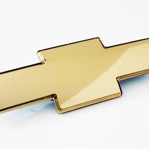 May include: A gold-colored Chevrolet emblem, featuring the iconic bowtie design. The emblem has a glossy finish and a beveled edge, reflecting light. The classic car part is set against a white background.