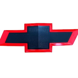 May include: A red and black Chevrolet emblem, featuring a black center with a textured surface and a red outline. The emblem is a stylized cross shape, commonly used as a car logo.