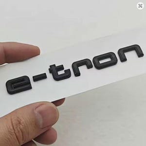 Könnte beinhalten: Schwarze, erhabene Buchstaben, die "e-tron" buchstabieren, auf weißem Hintergrund. Die Buchstaben sind dick und blockartig, mit leicht abgerundeter Optik. Das Bild wird von einer Hand gehalten.