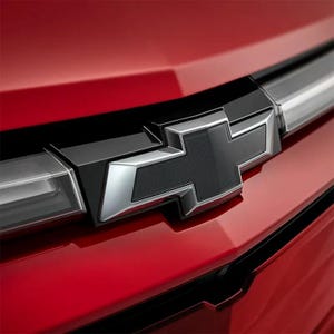 May include: Close-up of a red car's rear with a black and silver Chevrolet emblem. The emblem is a stylized cross with a textured black center and silver outline. The car's surface is glossy.