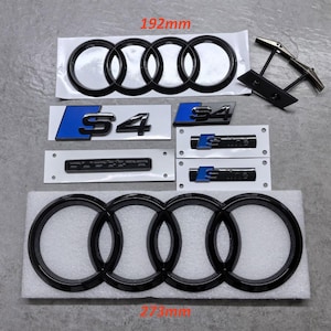 May include: A collection of black car emblems and badges, including the Audi rings, S4, and S-line logos. The largest emblem measures 273mm in length. Also included are a pair of black and silver cufflinks.