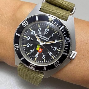 May include: A Swiss Raid Commando wristwatch with a black dial, white numerals, and a rotating black bezel. The watch features a green fabric strap, silver-tone case, and a small emblem with a yellow flame and a red and white shield on the face.