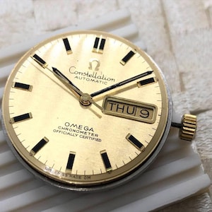 Puede incluir: Una esfera de reloj autom&aacute;tica Omega Constellation dorada con marcadores de hora y agujas rectangulares negros. La esfera del reloj muestra "Constellation Automatic" y "Omega Chronometer Officially Certified". La fecha es "JUE 9".