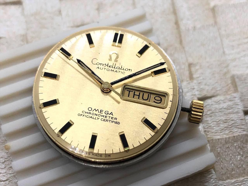 Puede incluir: Una esfera de reloj autom&aacute;tica Omega Constellation dorada con marcadores de hora y agujas rectangulares negros. La esfera del reloj muestra "Constellation Automatic" y "Omega Chronometer Officially Certified". La fecha es "JUE 9".