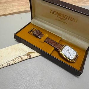 May include: A gold-tone Longines wristwatch with a white rectangular face, black Roman numeral markers, and a brown textured leather strap. The watch is presented in a brown hinged box with a gold-colored interior lining and the Longines logo.