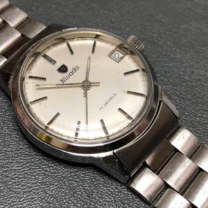 Puede incluir: Un reloj de pulsera Nivada vintage con una correa y caja de metal plateado. La esfera del reloj es blanca con marcadores negros, el logotipo de Nivada y una pantalla de fecha a las 3 en punto. Un reloj cl&aacute;sico.