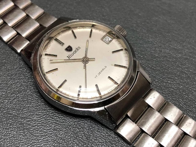 Puede incluir: Un reloj de pulsera Nivada vintage con una correa y caja de metal plateado. La esfera del reloj es blanca con marcadores negros, el logotipo de Nivada y una pantalla de fecha a las 3 en punto. Un reloj cl&aacute;sico.