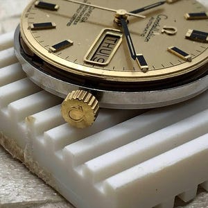 Puede incluir: Primer plano de un reloj autom&aacute;tico Omega Constellation dorado. La esfera muestra el d&iacute;a y la fecha, con marcadores de hora y agujas rectangulares negros. El reloj tiene una corona dorada con el logotipo de Omega y un bisel plateado. El reloj descansa sobre una superficie blanca.