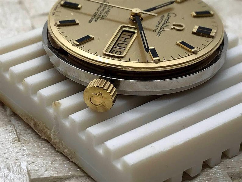 Puede incluir: Primer plano de un reloj autom&aacute;tico Omega Constellation dorado. La esfera muestra el d&iacute;a y la fecha, con marcadores de hora y agujas rectangulares negros. El reloj tiene una corona dorada con el logotipo de Omega y un bisel plateado. El reloj descansa sobre una superficie blanca.
