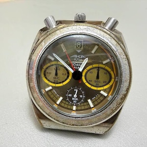 May include: A vintage AKA chronograph watch with a silver case and a dark brown face. The watch has yellow sub-dials and white hands. The text "AKA 1/10 CHRONOGRAPH OBA ALB" is visible. The watch has a textured, aged appearance.