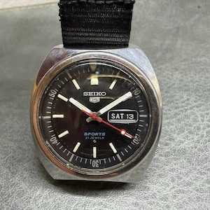 May include: A Seiko 5 Sports wristwatch with a black dial, silver-tone case, and black nylon strap. The dial features luminous hour markers, a day-date window displaying SAT 13, a red second hand, and text reading SEIKO, 5, SPORTS, and 21 JEWELS.
