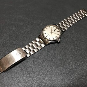Puede incluir: Un reloj de pulsera plateado con esfera blanca y correa plateada. El reloj tiene una esfera redonda con agujas e indicadores plateados. La correa est&aacute; hecha de peque&ntilde;os eslabones plateados rectangulares. El cierre tiene un logotipo.