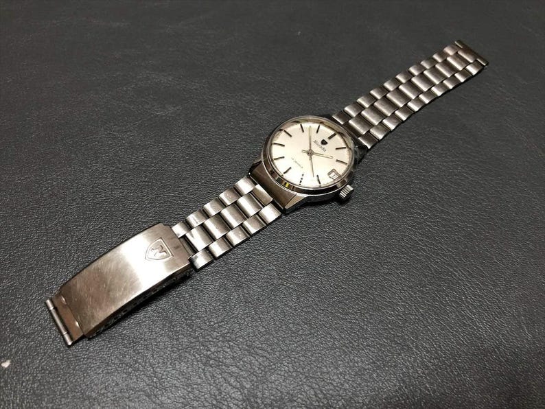 Puede incluir: Un reloj de pulsera plateado con esfera blanca y correa plateada. El reloj tiene una esfera redonda con agujas e indicadores plateados. La correa est&aacute; hecha de peque&ntilde;os eslabones plateados rectangulares. El cierre tiene un logotipo.