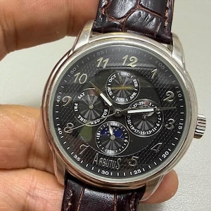 May include: A silver Arbutus watch with a black face, featuring subdials for day, date, and moon phase. It has a brown leather strap and a textured dial. The watch face displays the numbers 1 to 12.