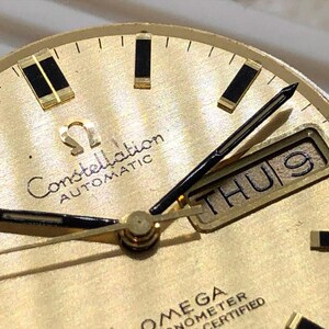 Puede incluir: Primer plano de una esfera de reloj autom&aacute;tico Omega Constellation dorada. El reloj presenta agujas e indicadores negros, una pantalla de d&iacute;a y fecha que muestra "THU 9" y el logotipo de Omega. La esfera del reloj tiene un acabado texturizado.