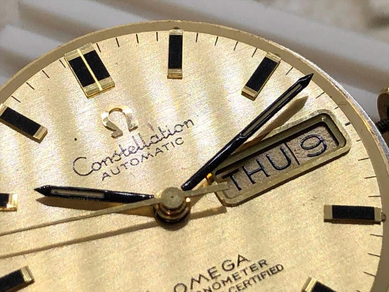 Puede incluir: Primer plano de una esfera de reloj autom&aacute;tico Omega Constellation dorada. El reloj presenta agujas e indicadores negros, una pantalla de d&iacute;a y fecha que muestra "THU 9" y el logotipo de Omega. La esfera del reloj tiene un acabado texturizado.