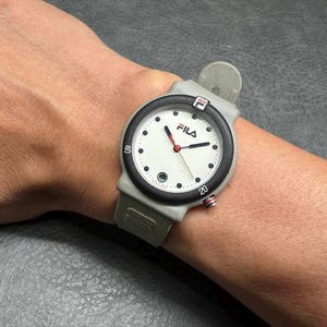 May include: A Fila watch with a white face, black bezel, and gray band is shown on a wrist. The watch has red hands and black dot hour markers. The Fila logo is visible on the watch face.