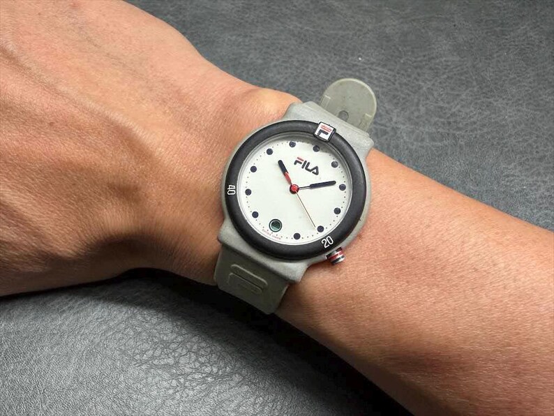 May include: A Fila watch with a white face, black bezel, and gray band is shown on a wrist. The watch has red hands and black dot hour markers. The Fila logo is visible on the watch face.