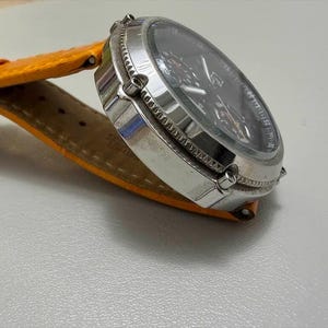 以下が含まれることがあります： 黒い文字盤とオレンジ色の革バンドが付いた、シルバーの腕時計。 時計は、複数のダイヤルとテクスチャード加工されたベゼルを備えた丸い文字盤です。 バンドは明るいオレンジ色で、革製のように見えます。