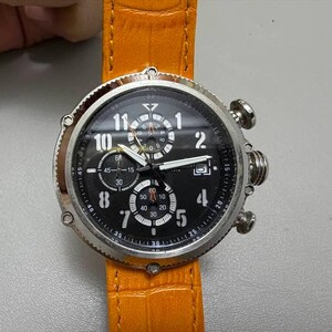 以下が含まれることがあります： 黒い文字盤と白い数字が付いたシルバーの腕時計。オレンジ色の革バンドが付いています。文字盤には複数のサブダイヤルと針が表示され、1から12までの数字が見えます。