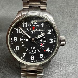 May include: A stainless steel Uir-Kraft wristwatch with a black dial, white Arabic numerals, and a date window at the three o'clock position. The watch features a GMT function with a red-tipped hand, luminous hands, and a metal link bracelet.