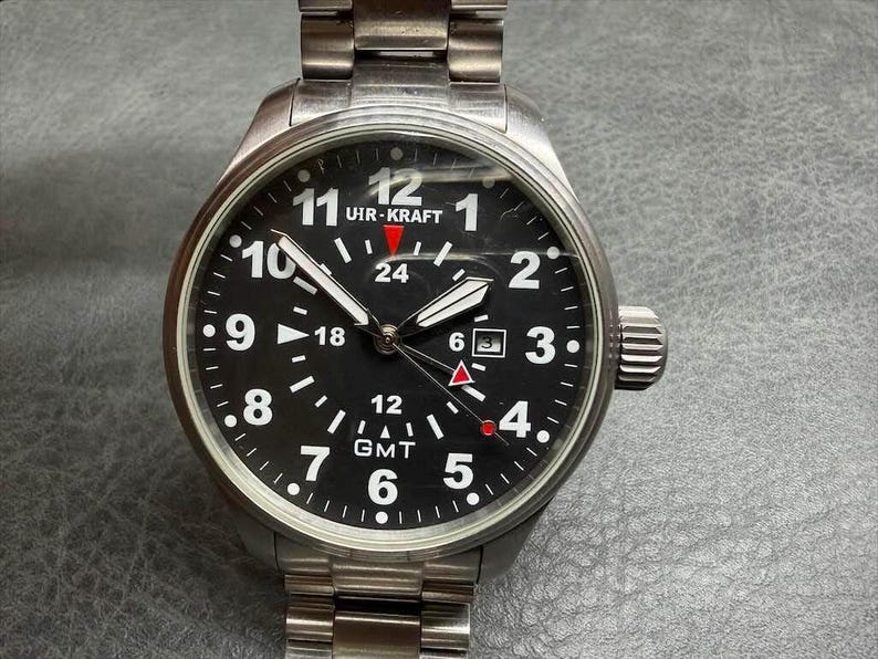 May include: A stainless steel Uir-Kraft wristwatch with a black dial, white Arabic numerals, and a date window at the three o'clock position. The watch features a GMT function with a red-tipped hand, luminous hands, and a metal link bracelet.