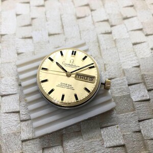Puede incluir: Una esfera de reloj autom&aacute;tica Omega Constellation dorada con marcadores de hora y agujas negras. La esfera muestra el d&iacute;a y la fecha. El texto "Constellation Automatic" y "Omega Chronometer Officially Certified" es visible.