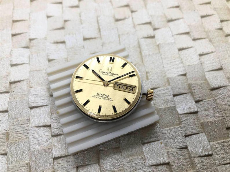 Puede incluir: Una esfera de reloj autom&aacute;tica Omega Constellation dorada con marcadores de hora y agujas negras. La esfera muestra el d&iacute;a y la fecha. El texto "Constellation Automatic" y "Omega Chronometer Officially Certified" es visible.